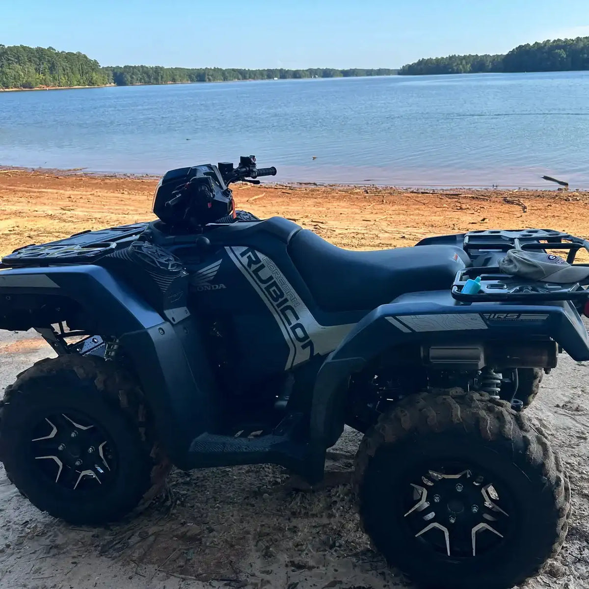 Black honda rubicon atv with rugged tires and a matte finish, parked on sandy shore near water.