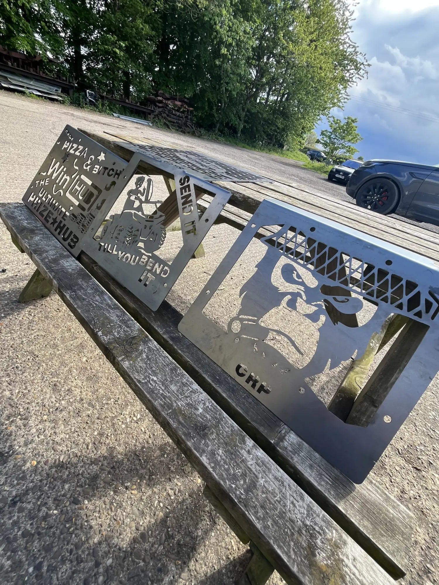 Metallic gray steel cutout signs featuring bold, edgy designs including a gorilla face and humorous text like ’pizza & bitch’ and ’send it,’ displayed on a wooden bench.